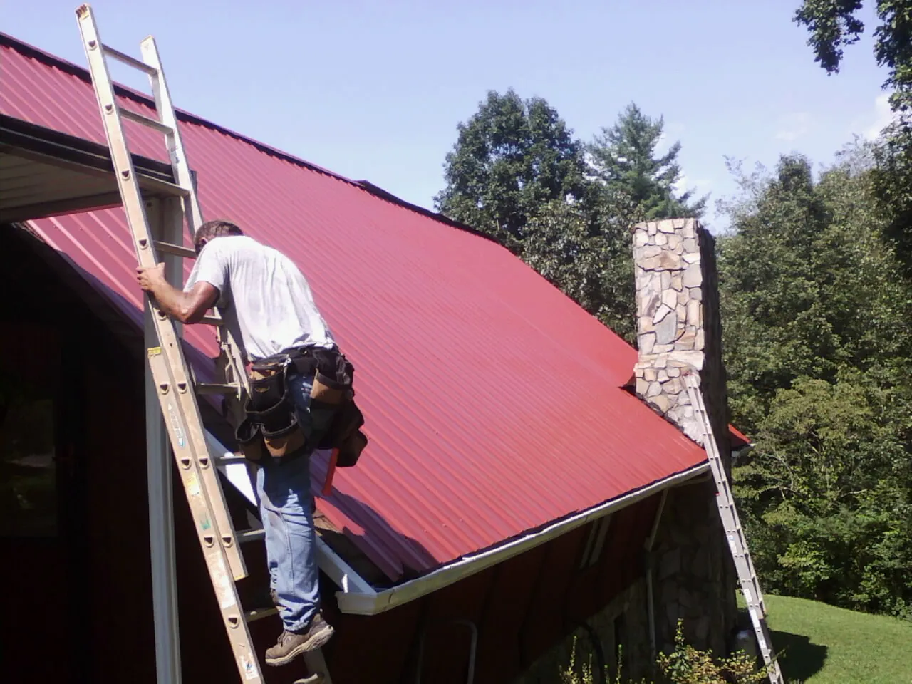 Roofer on a ladder beside a red metal roof and stone chimney — Soffit & Fascia Repair by Roofing Brothers in Mooseheart